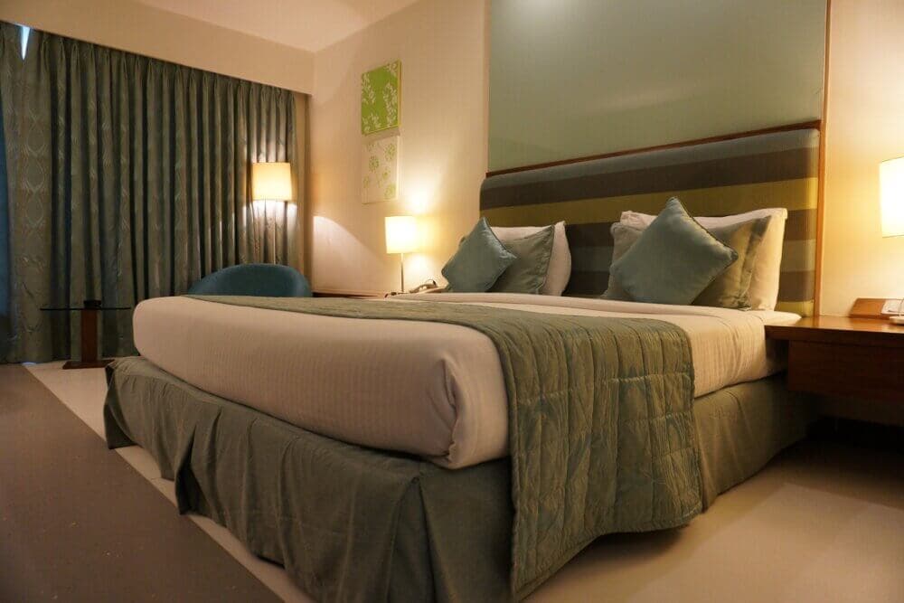 A cozy hotel room with a neatly made double bed, soft lighting, curtains, and a side table with a lamp. - Home Instead
