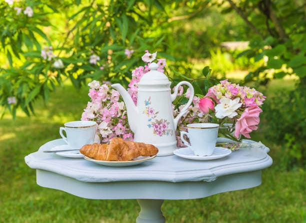 Elegant garden table with floral teapot, two teacups, croissants, and fresh flower bouquet on a sunny day. - Home Instead