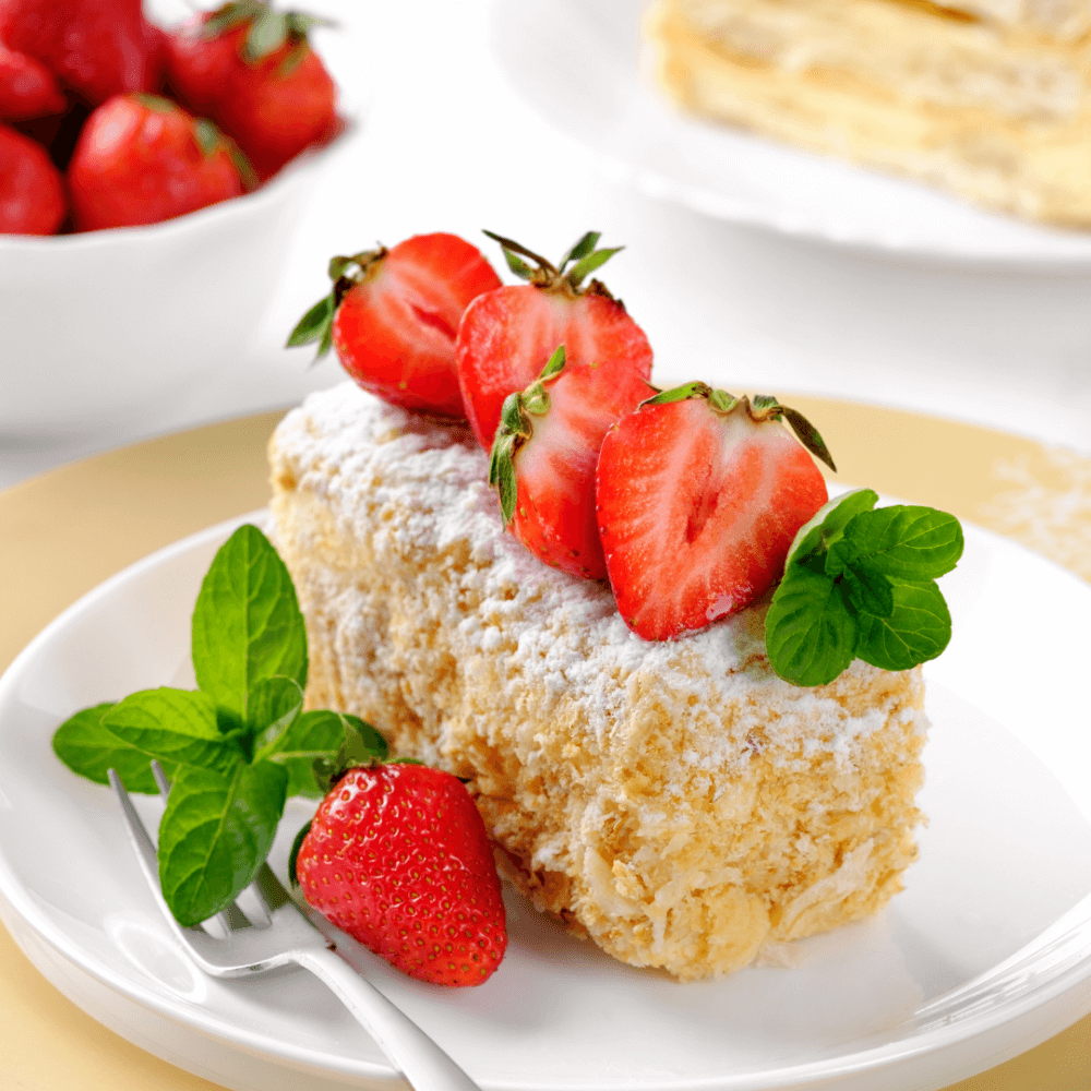 A slice of cake topped with powdered sugar, fresh strawberries, and mint, with more strawberries on the side. - Home Instead