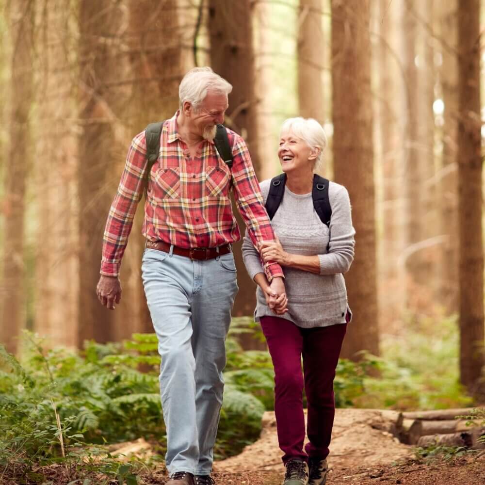 Elderly couple walking hand-in-hand on a forest trail, smiling and enjoying a peaceful moment together. - Home Instead