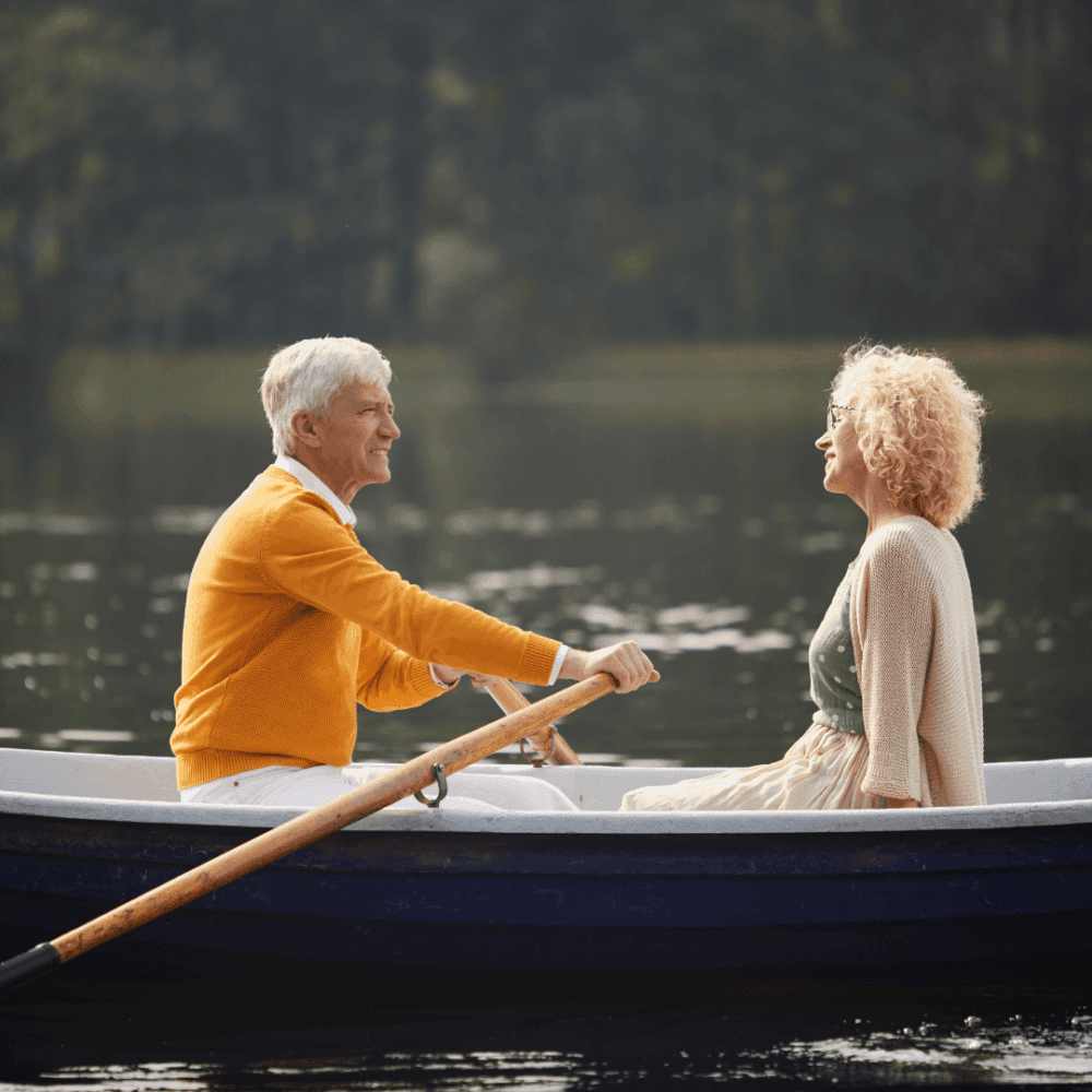 An elderly man rows a woman in a boat on a calm lake; both are smiling and enjoying the serene environment. - Home Instead