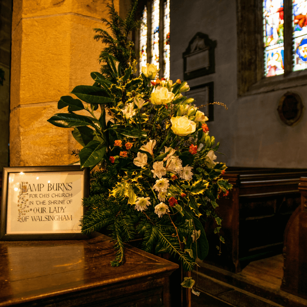 A flower arrangement with a sign reading "LAMP BURNS FOR THIS CHURCH IN THE SHRINE OF OUR LADY OF WALSINGHAM" in a church. - Home Instead