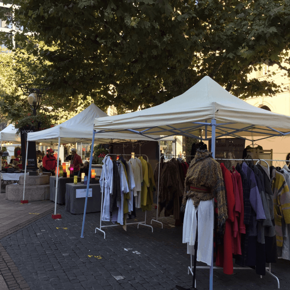 Outdoor market with white tents displaying clothes on racks, vendors, and shoppers under leafy trees. - Home Instead