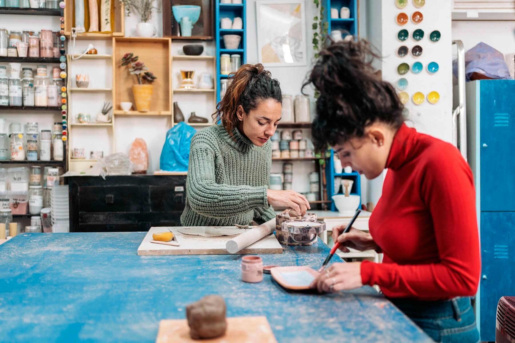 Two women working on pottery projects at a blue table in a studio filled with ceramics and art supplies. - Home Instead