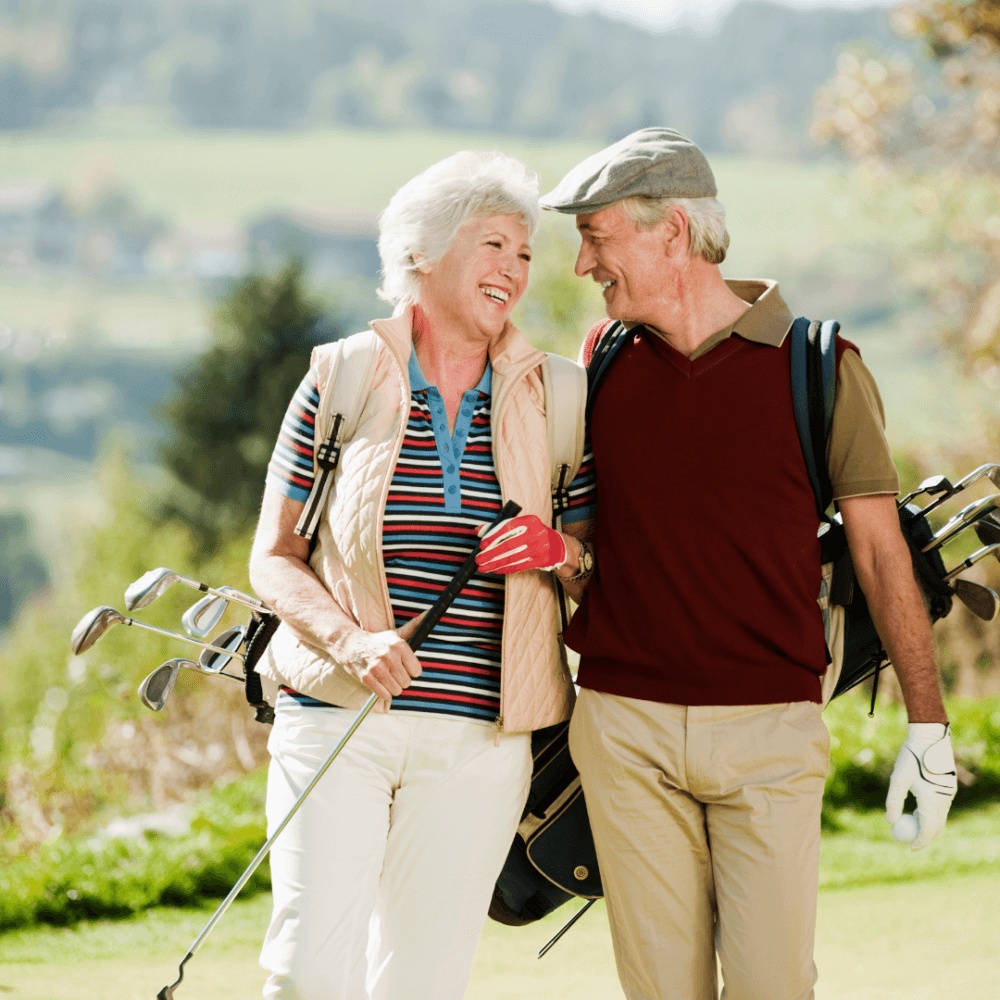 An elderly couple with golf clubs smiles and walks on a scenic golf course. - Home Instead