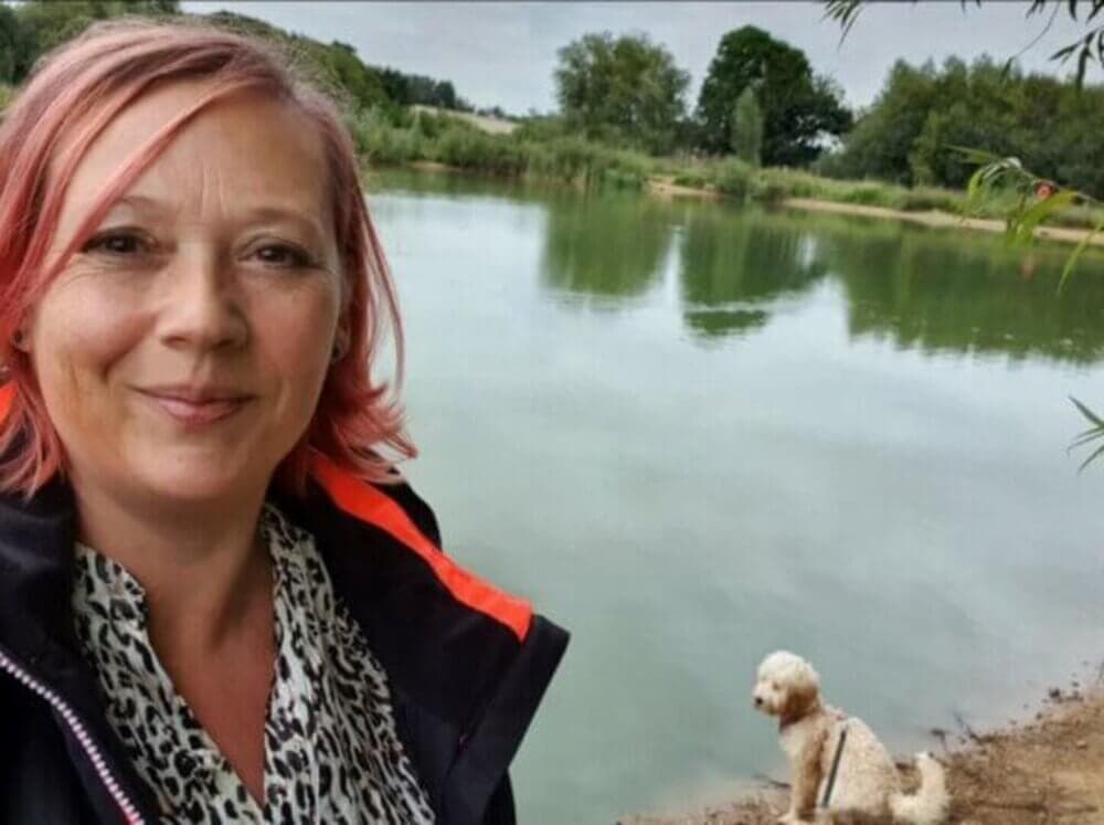 Woman with pink hair smiles in front of a serene lake, with a small white dog near the water's edge. - Home Instead