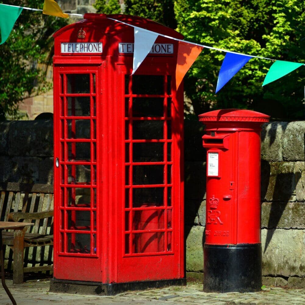 A red British telephone box and post box are adorned with colorful flags, with a stone wall and greenery in the background. - Home Instead