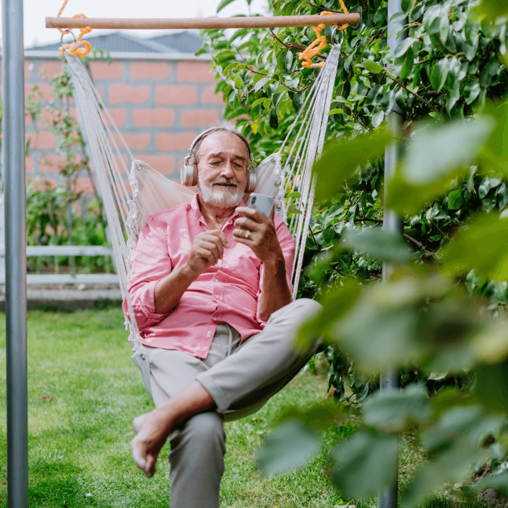 A smiling man in a pink shirt and headphones relaxes in a hammock, using his phone in a garden. - Home Instead