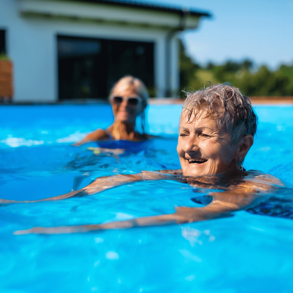Two elderly women enjoying a swim in a bright blue outdoor pool with a house in the background. - Home Instead