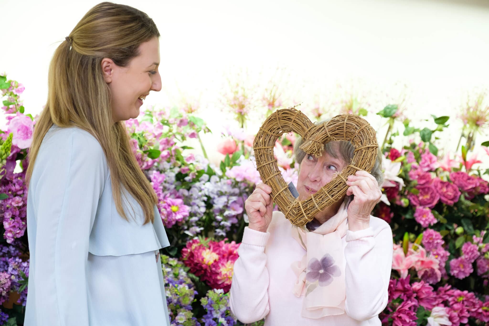 Two women sharing a laugh, one holding a heart-shaped wicker frame in front of her face, against a floral backdrop. - Home Instead