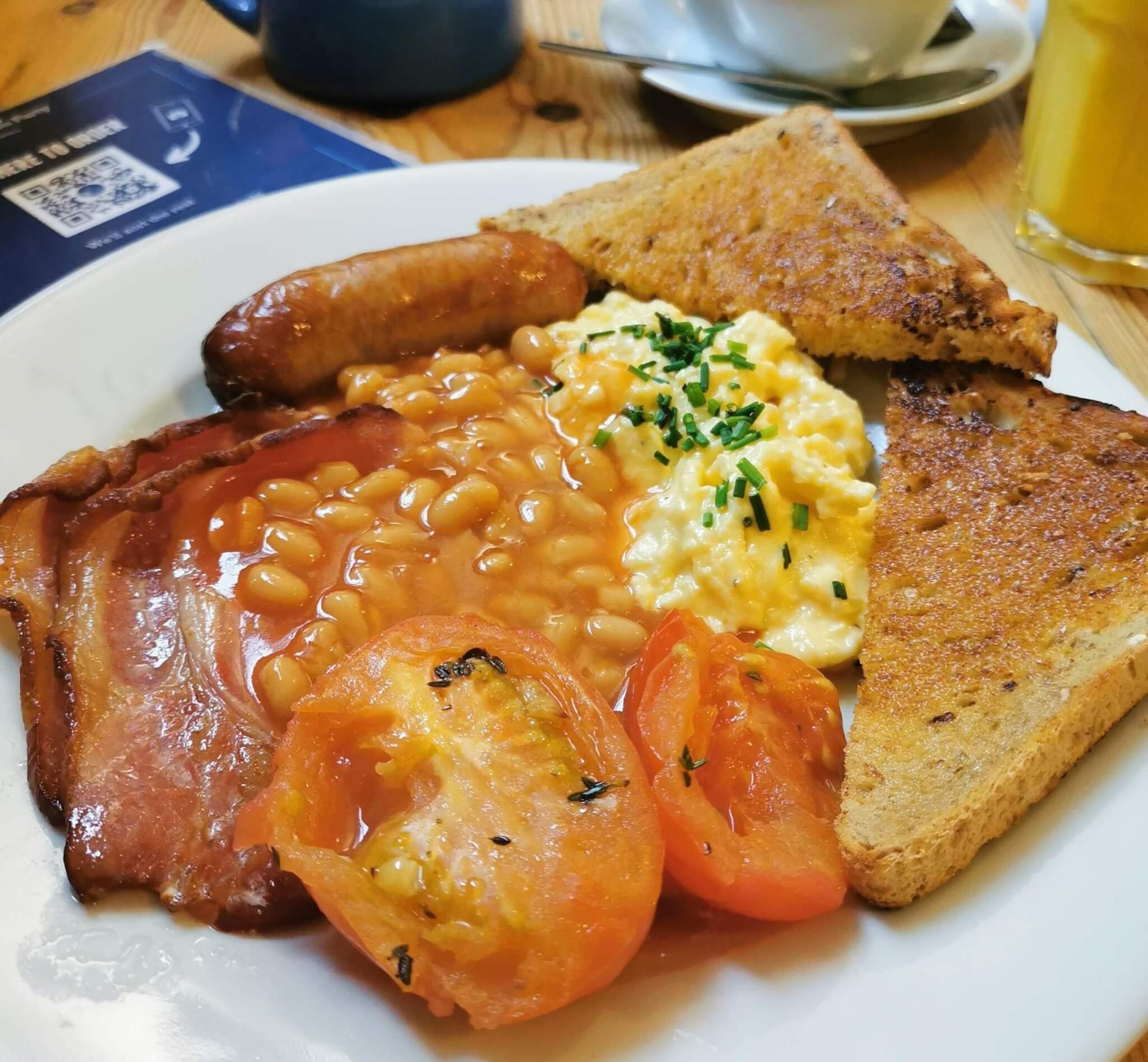 A full English breakfast with bacon, sausage, baked beans, scrambled eggs, grilled tomatoes, and toast on a white plate. - Home Instead