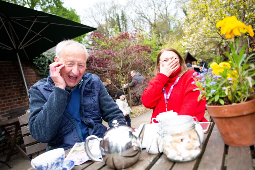 An elderly man and a young woman laugh joyfully at an outdoor café table with tea in front of them. - Home Instead