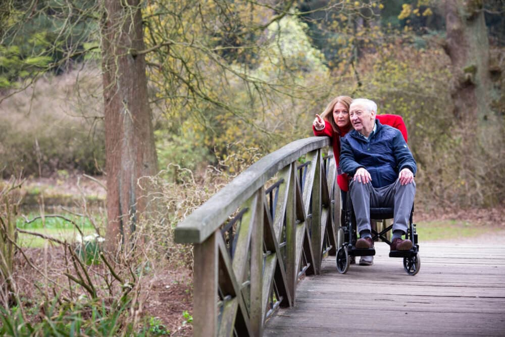 A smiling elderly man in a wheelchair and a woman pointing while on a wooden bridge in a nature park. - Home Instead