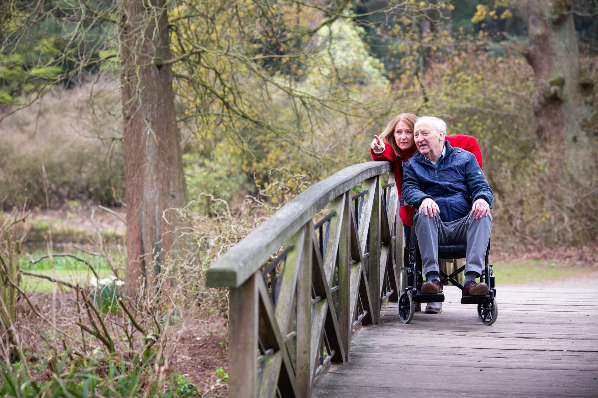 A woman assists an elderly man in a wheelchair across a wooden bridge in a lush park setting. - Home Instead