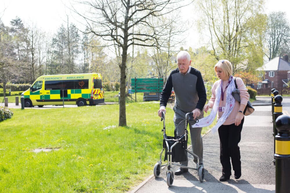 Elderly man using a walker with assistance from a caregiver outside, with an ambulance parked in the background. - Home Instead