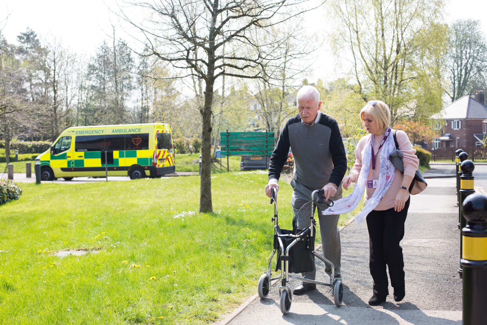 Elderly man using a walker with assistance from a caregiver outside, with an ambulance parked in the background. - Home Instead