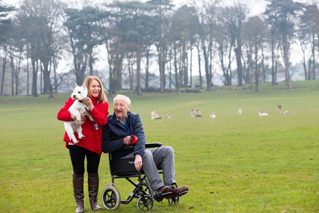 A woman holds a white dog while standing next to a man in a wheelchair in a field with deer and trees in the background. - Home Instead
