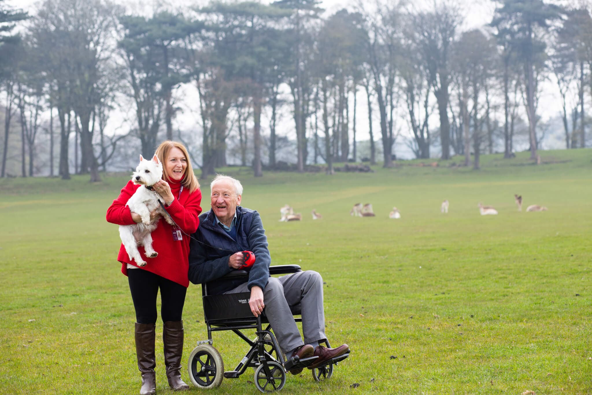 A woman in a red jacket holds a dog while standing next to an elderly man in a wheelchair in a grassy field with deer. - Home Instead