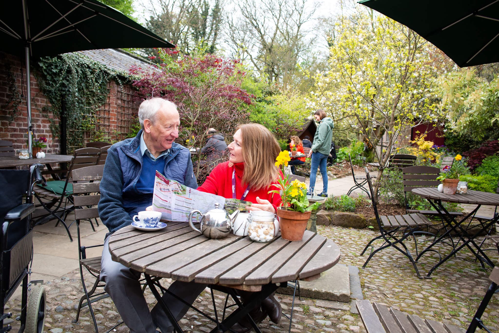An elderly man and a woman smile while seated at a cafe table outdoors, surrounded by lush greenery and flowers. - Home Instead