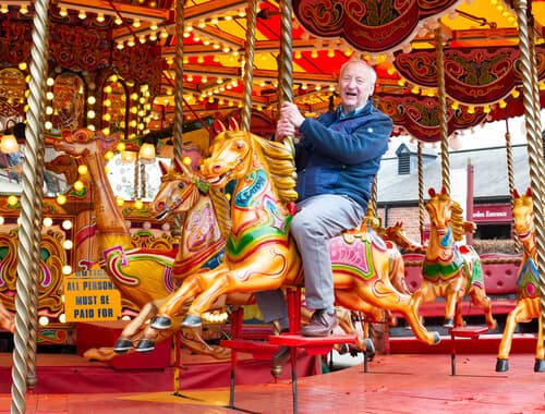 A joyful man riding a colorful, ornate carousel horse at a fairground with lights and decorations in the background. - Home Instead