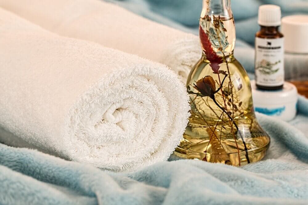 Rolled white towels and a glass bottle with flowers on a blue fuzzy blanket, with essential oil bottles in the background. - Home Instead