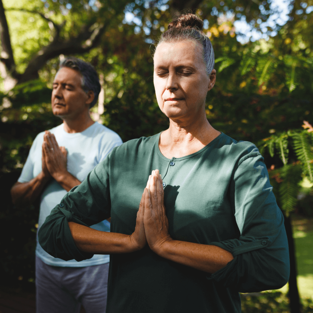 Two people meditating outside in nature, hands in prayer position, eyes closed, peaceful expressions. - Home Instead