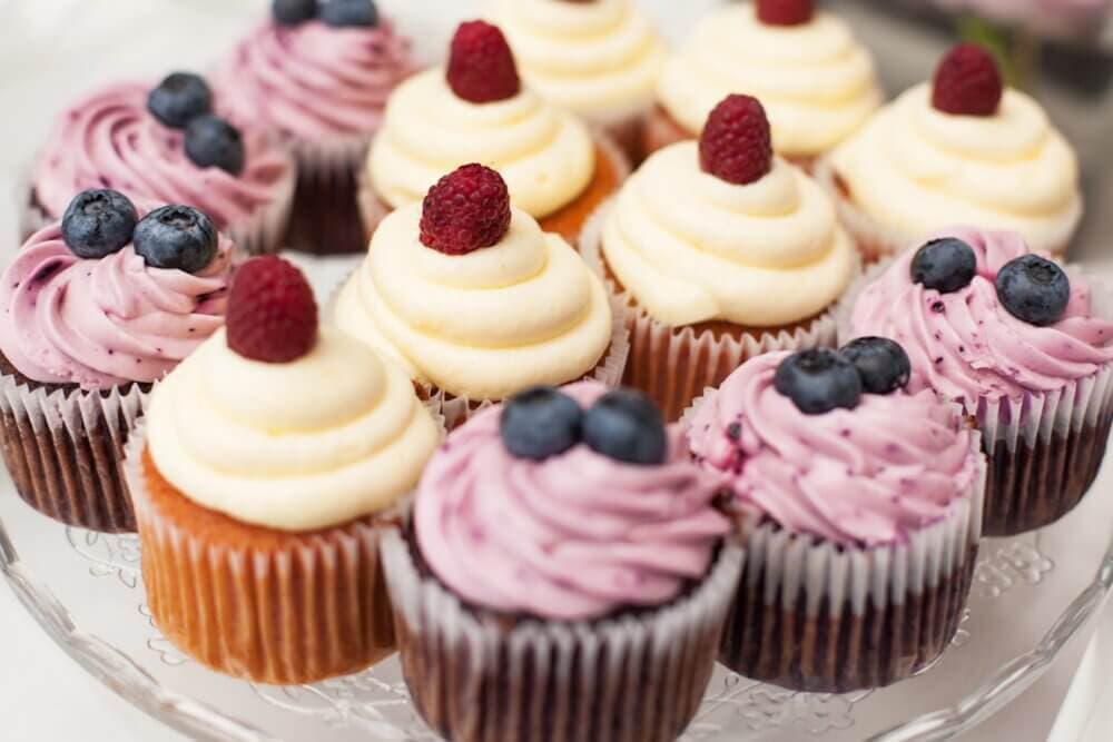 Cupcakes with white and purple frosting, topped with fresh raspberries and blueberries, arranged on a glass platter. - Home Instead