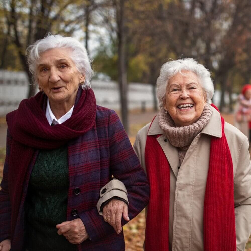 Two elderly women smiling and walking arm in arm outdoors during autumn. They are wearing coats and scarves. - Home Instead