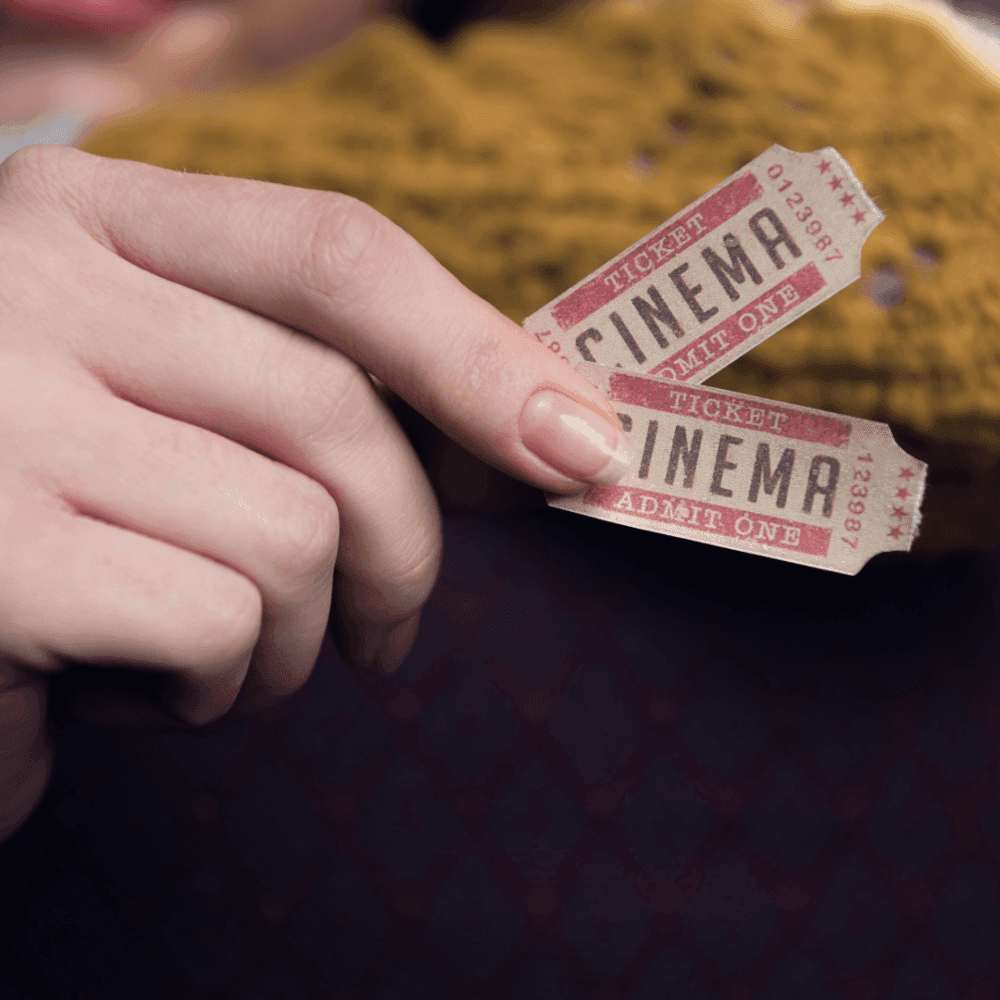 A hand holding two vintage cinema tickets with "Admit One" text. Polka-dot sleeve and yellow scarf in background. - Home Instead