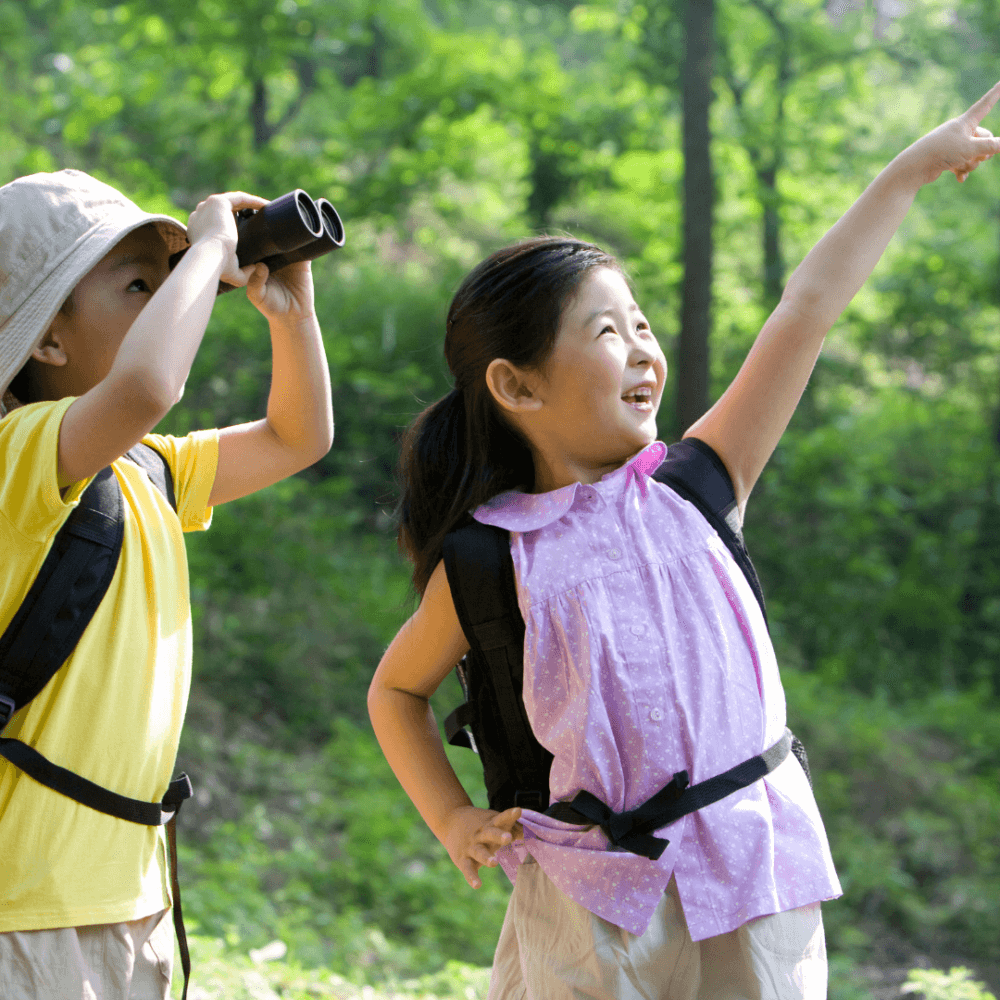 Two children with backpacks in a forest; one uses binoculars while the other points excitedly upward. - Home Instead