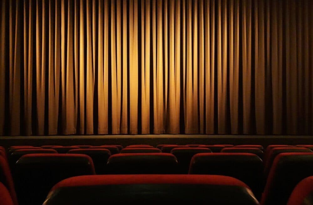 Empty theater with red and black seats facing a closed, beige stage curtain. - Home Instead