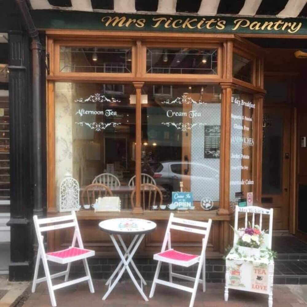 A quaint café with a sign "Mrs Tickit's Pantry," featuring a small outdoor seating area with two chairs and a table. - Home Instead