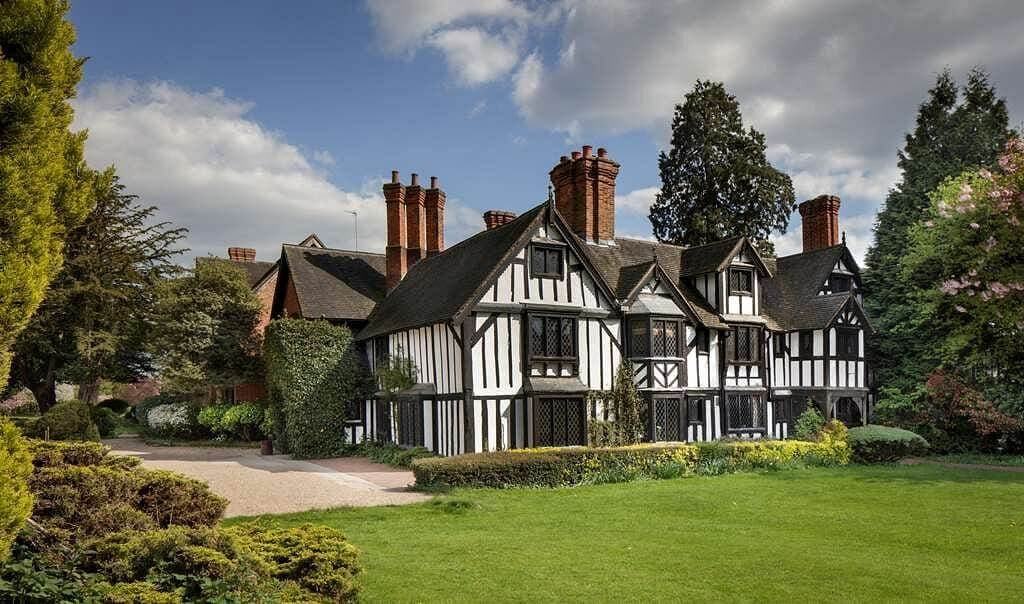 A historic black and white timber-framed house with multiple chimneys and a lush green garden on a sunny day. - Home Instead