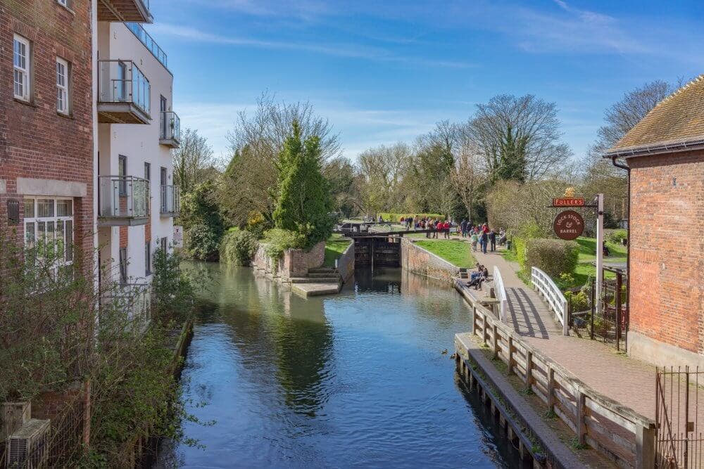 River Lock with adjacent buildings, trees, and people walking on the path; clear sky and calm water. - Home Instead