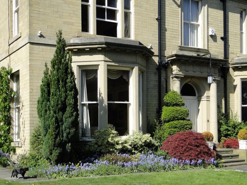 A brick house with a bay window and a well-tended garden featuring flowers, shrubs, and a black cat on the lawn. - Home Instead