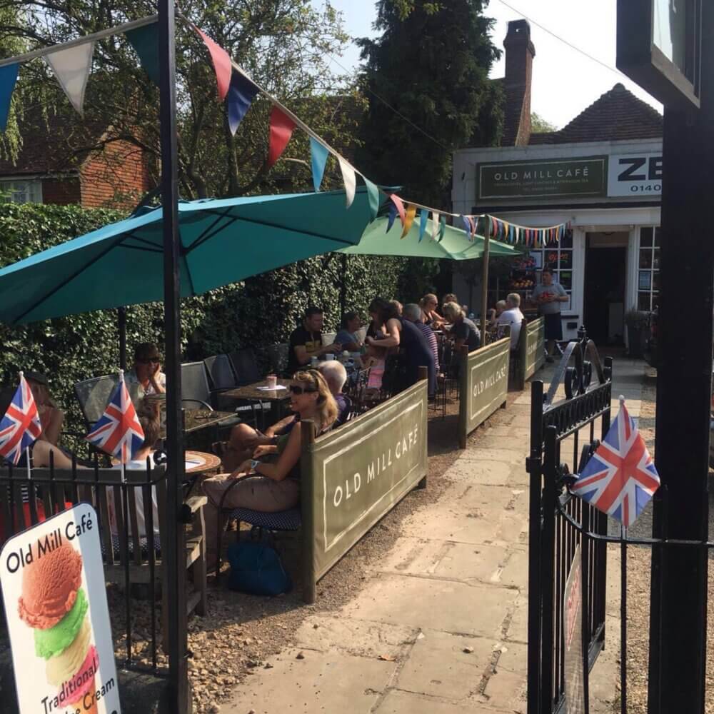 Outdoor seating at the Old Mill Cafe with patrons dining under umbrellas and bunting flags on a sunny day. - Home Instead
