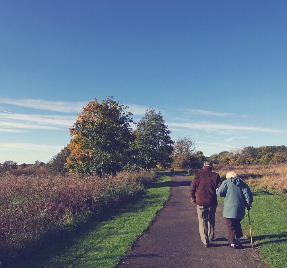 An elderly couple walks down a paved path surrounded by autumn trees and grass under a clear blue sky. - Home Instead