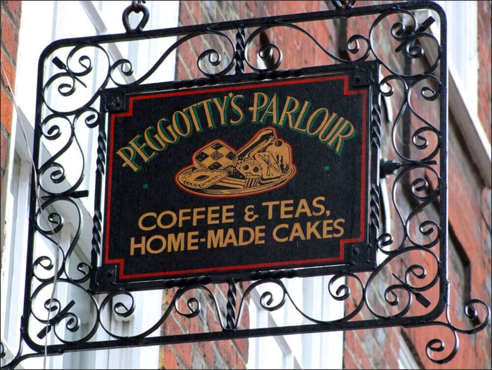 A hanging black sign reads "Peggotty's Parlour, Coffee & Teas, Home-Made Cakes" with an image of tea and cake. - Home Instead