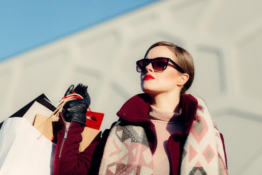 Woman in sunglasses holding shopping bags, wearing a scarf and gloves, with a geometric background. - Home Instead