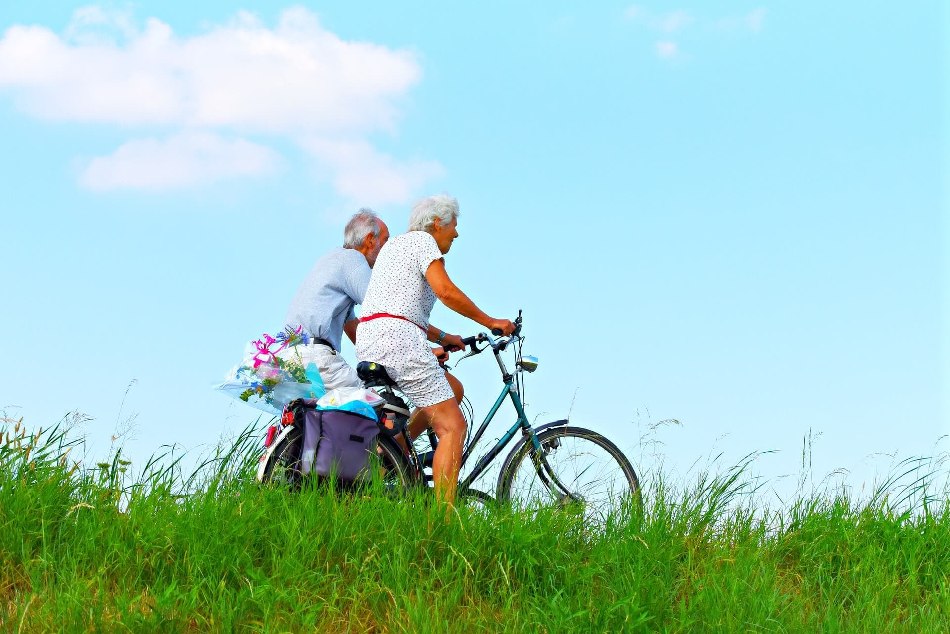 Two elderly people riding bicycles on a grassy path, with a blue sky and few clouds in the background. - Home Instead