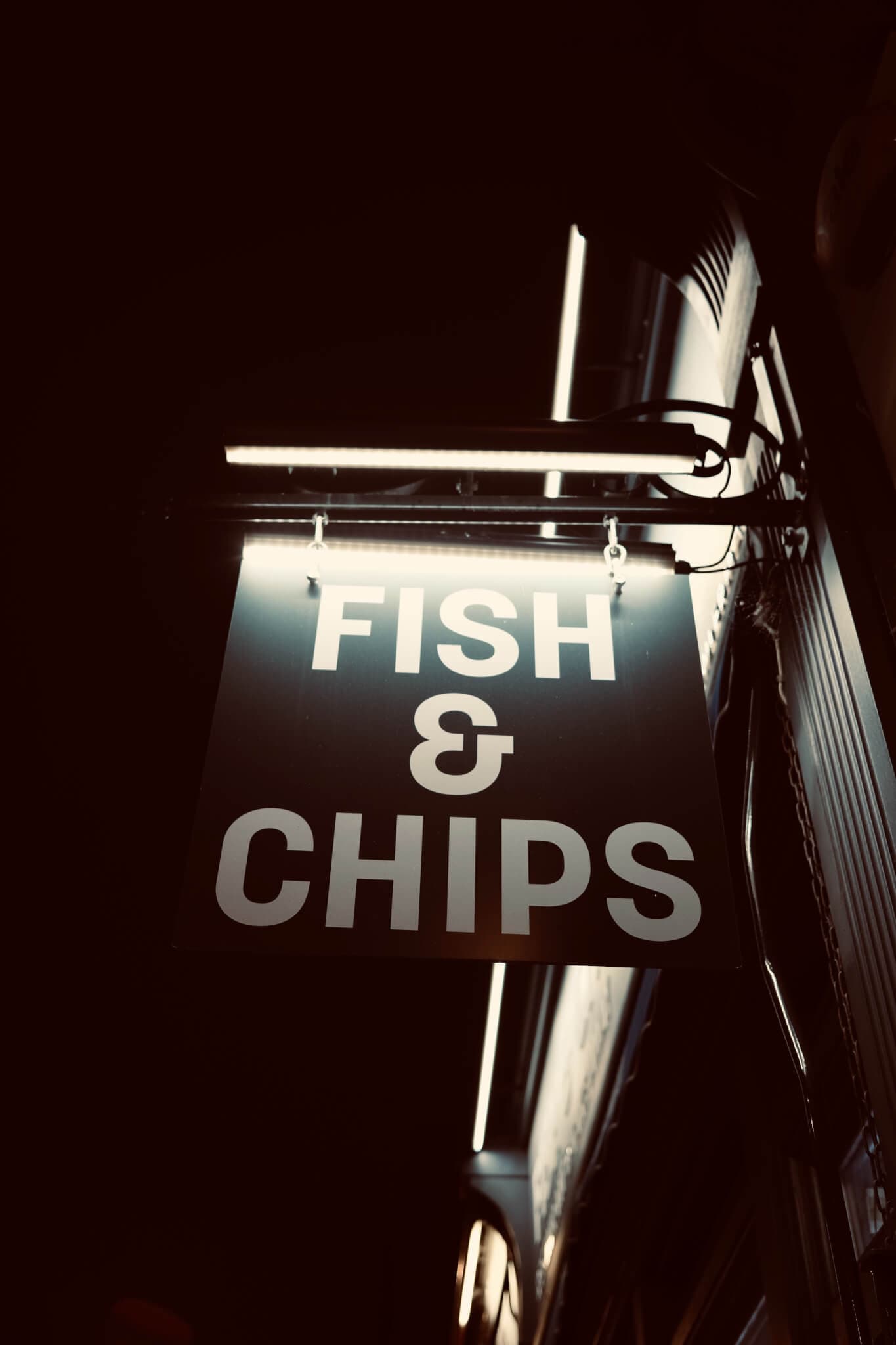 A hanging sign illuminated at night displaying "FISH & CHIPS" in bold white letters. - Home Instead