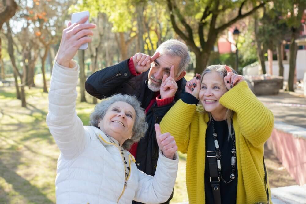 Three elderly people taking a playful selfie in a park on a sunny day, making fun gestures with their hands. - Home Instead