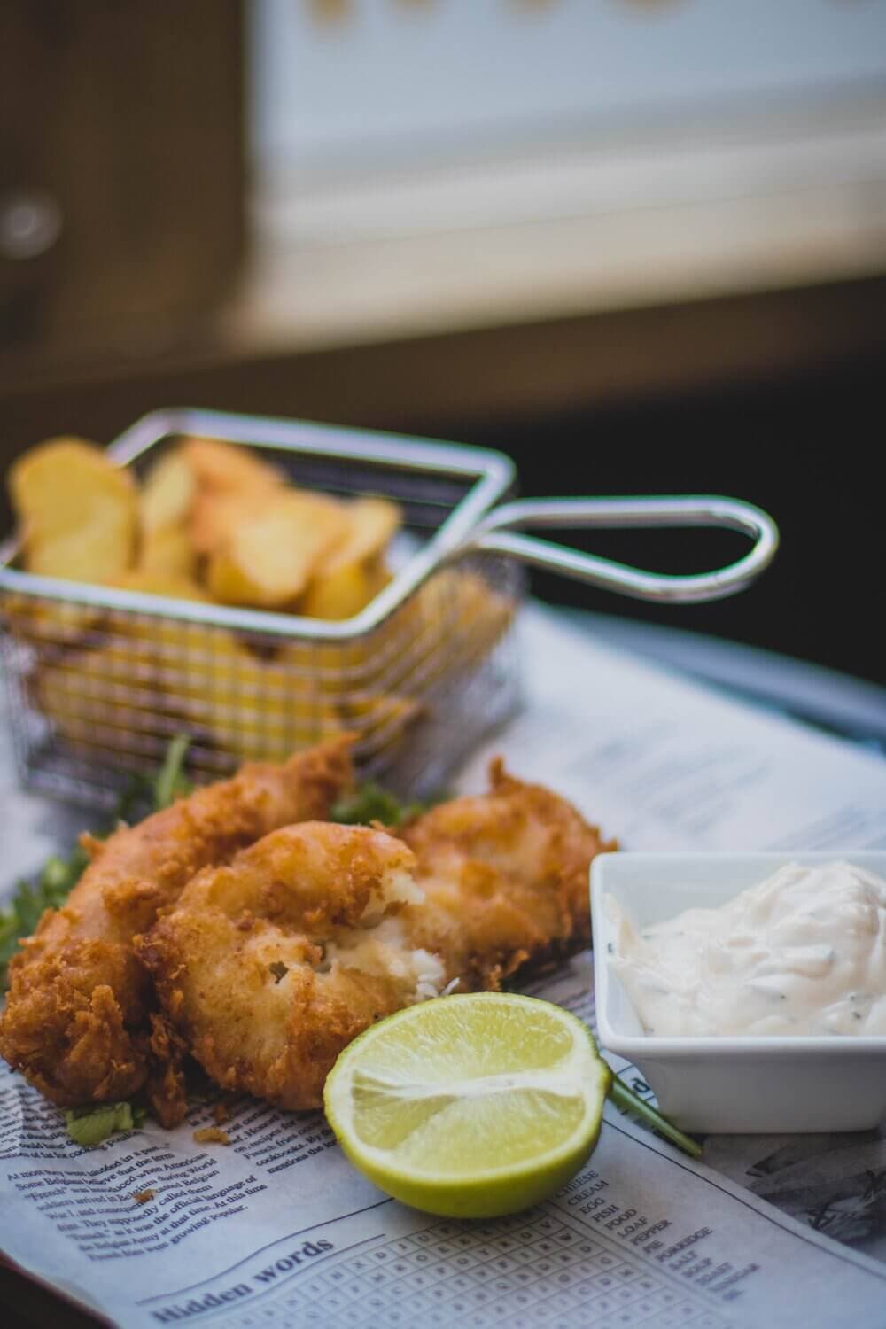 Fried fish with tartar sauce, a lime wedge, and a basket of fries on a sheet of newspaper. - Home Instead