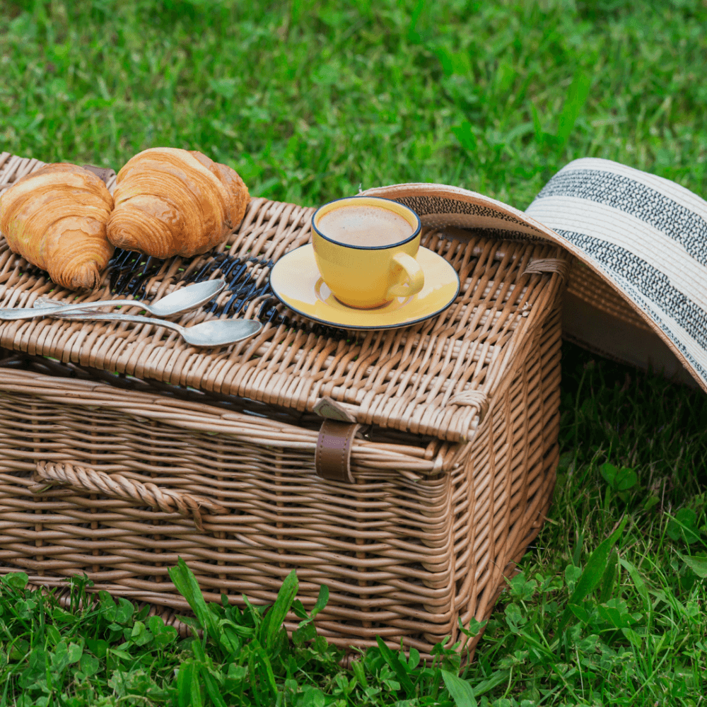 Wicker picnic basket with croissants, a cup of coffee in a yellow saucer, and a wide-brimmed hat on grass. - Home Instead