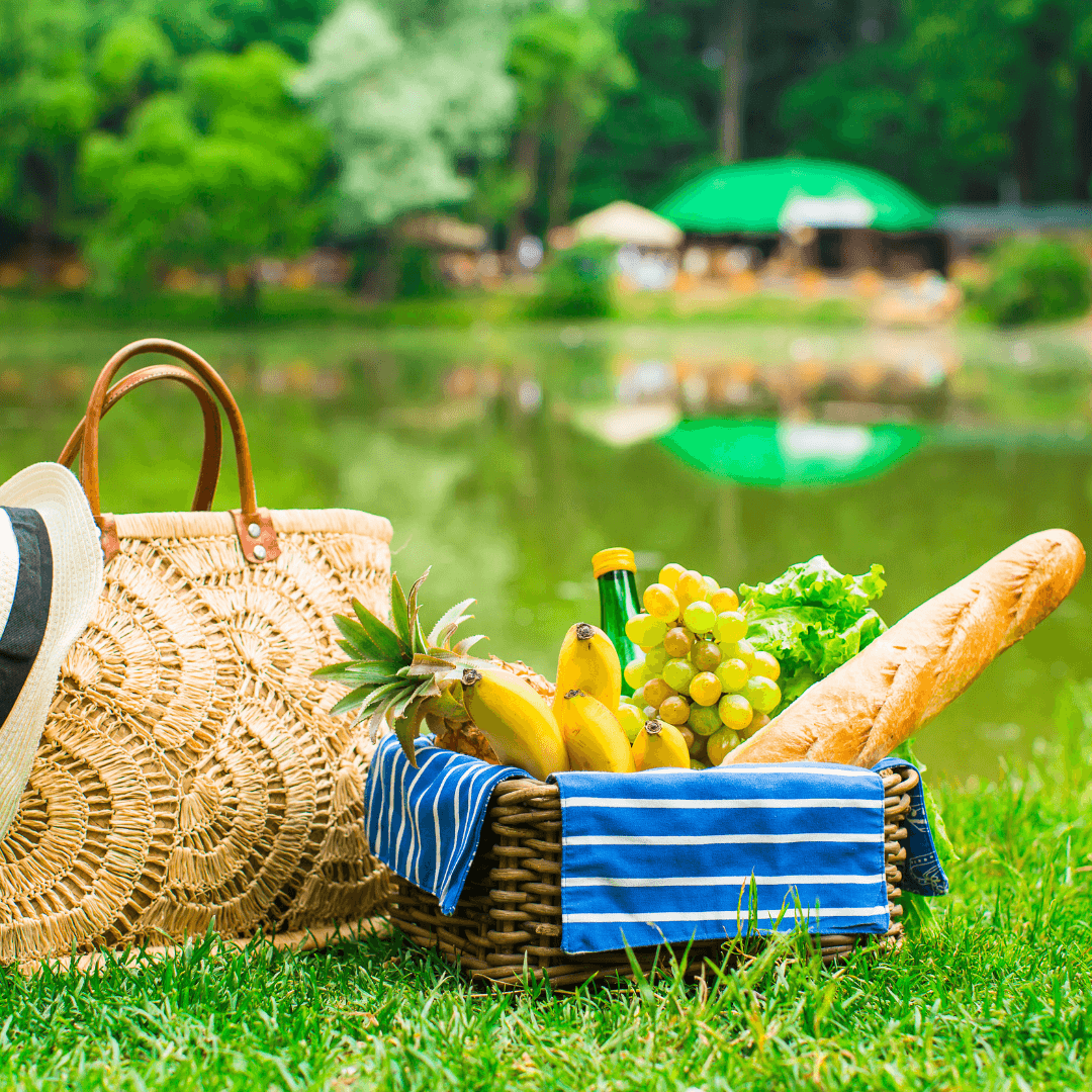 A picnic basket with fruits, a baguette, and a green bottle sits on grass near a tranquil lake, with a straw bag and hat beside it. - Home Instead