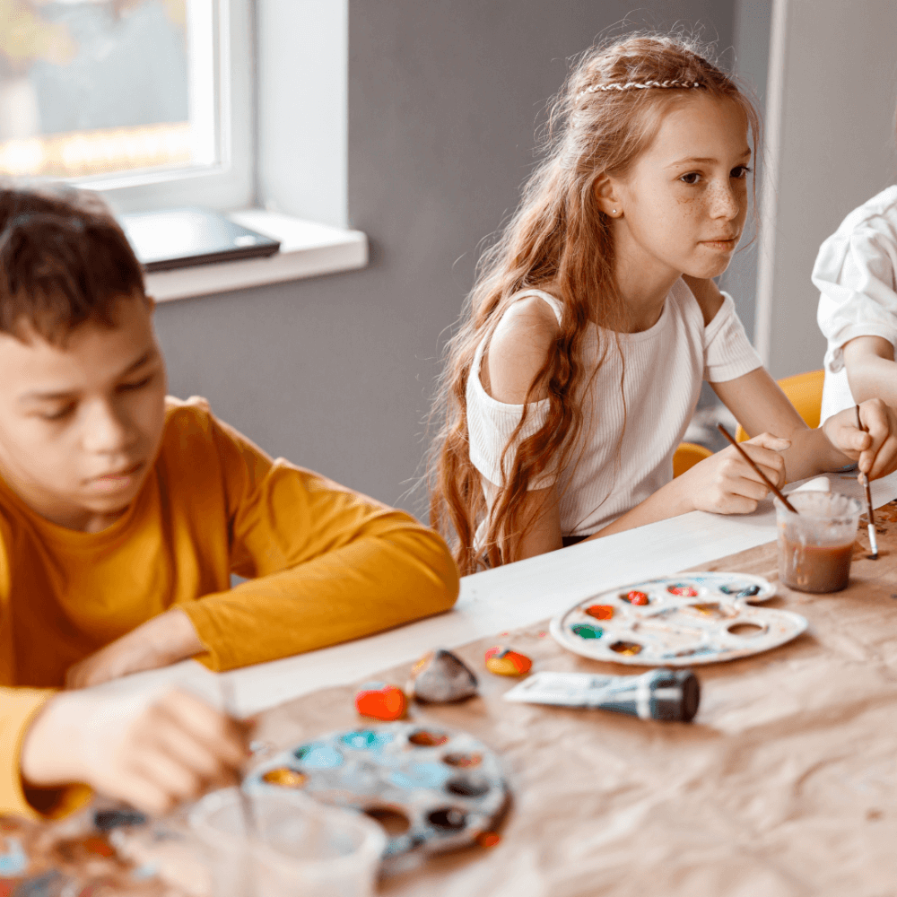 Three children painting at a table with brushes and palettes, focused on their artwork in a well-lit room. - Home Instead