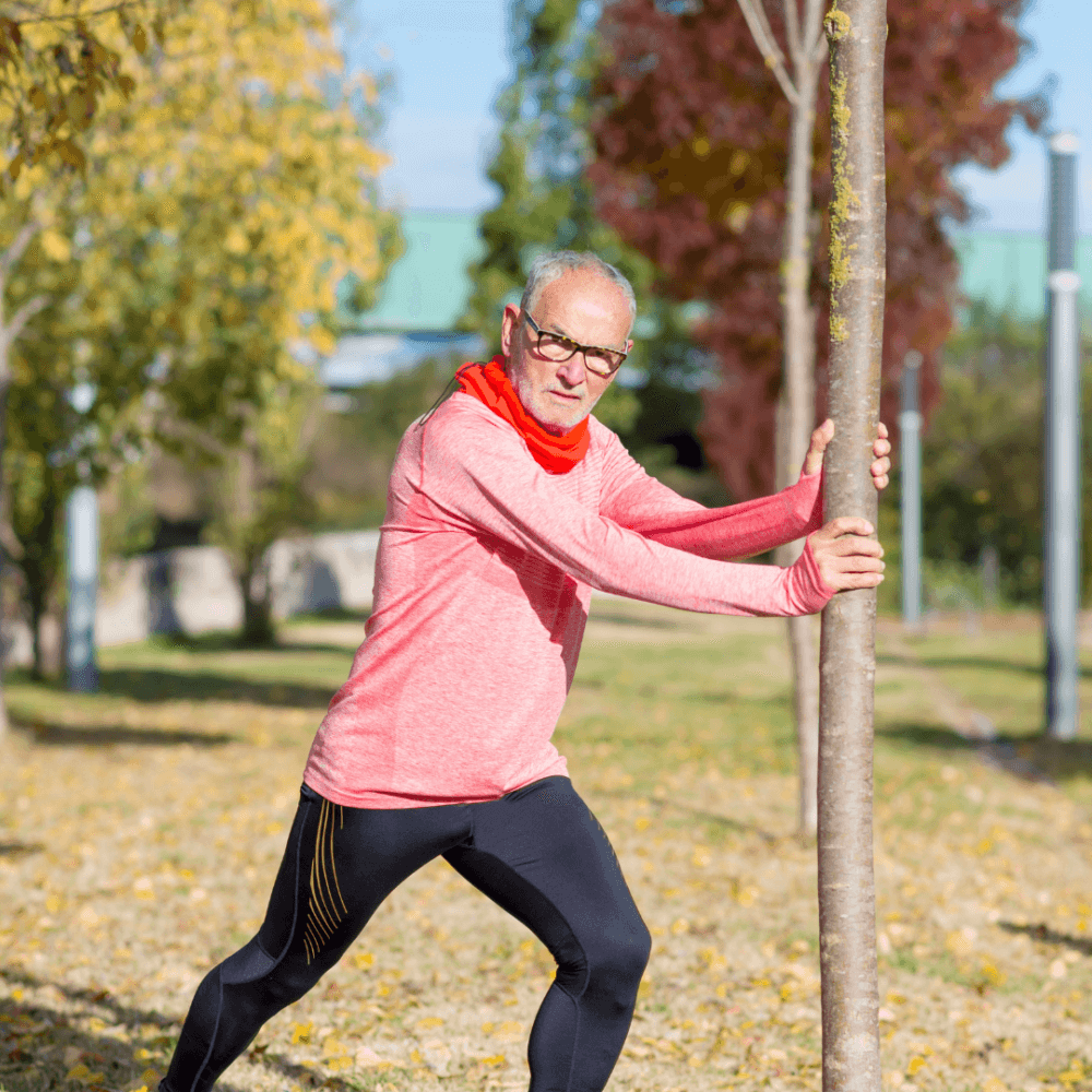 Older man stretching using a tree in a park, wearing glasses, red shirt, and black leggings on a sunny day. - Home Instead