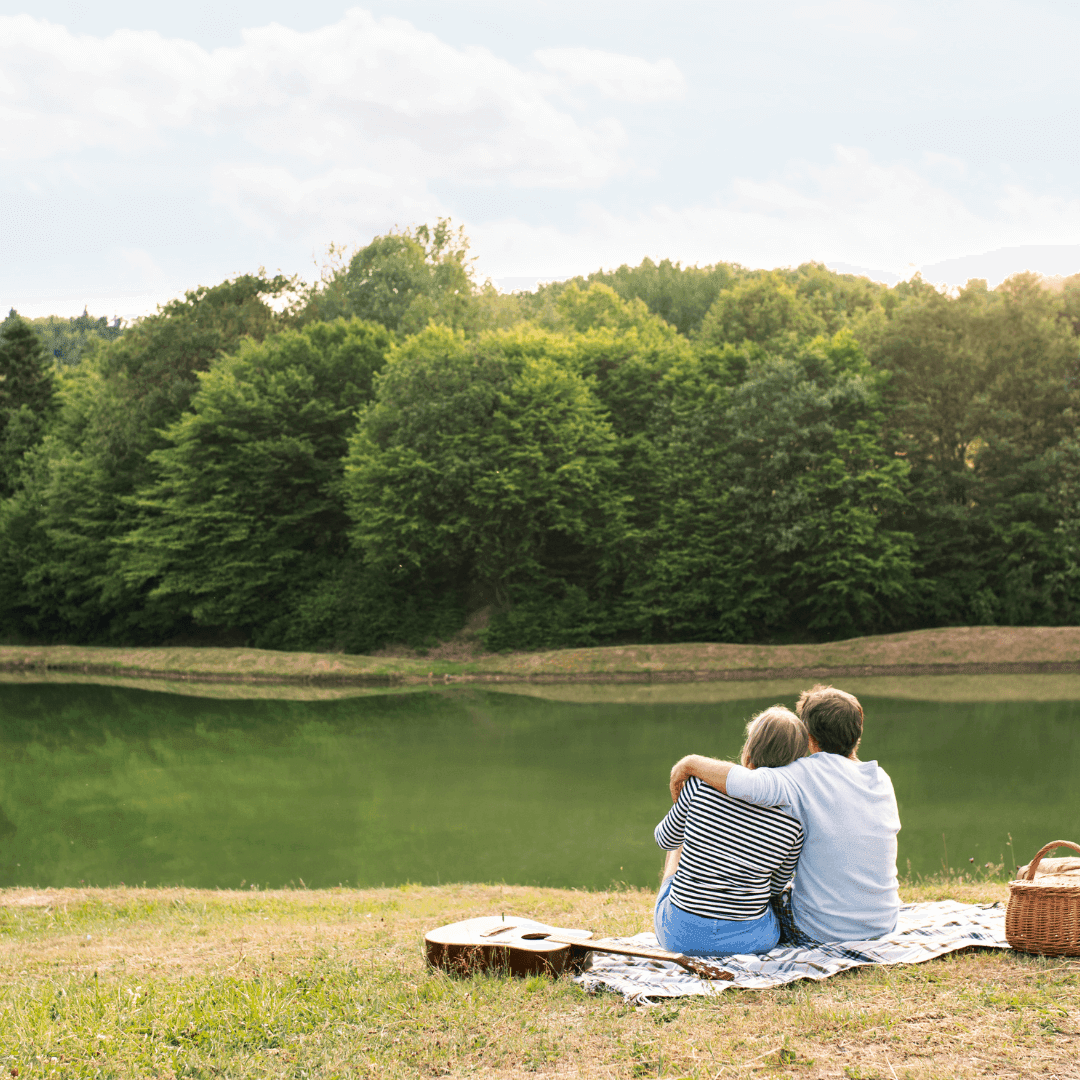 Two people sit on a blanket by a lake with a guitar and picnic basket, surrounded by trees on a sunny day. - Home Instead