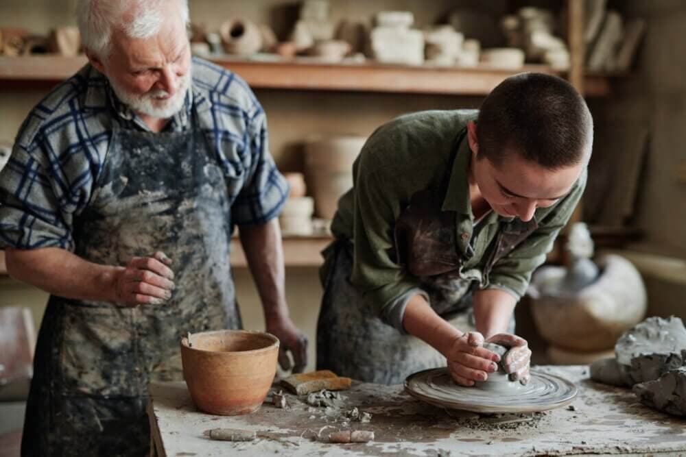 An older man guides a younger person shaping clay on a pottery wheel in a workshop. - Home Instead