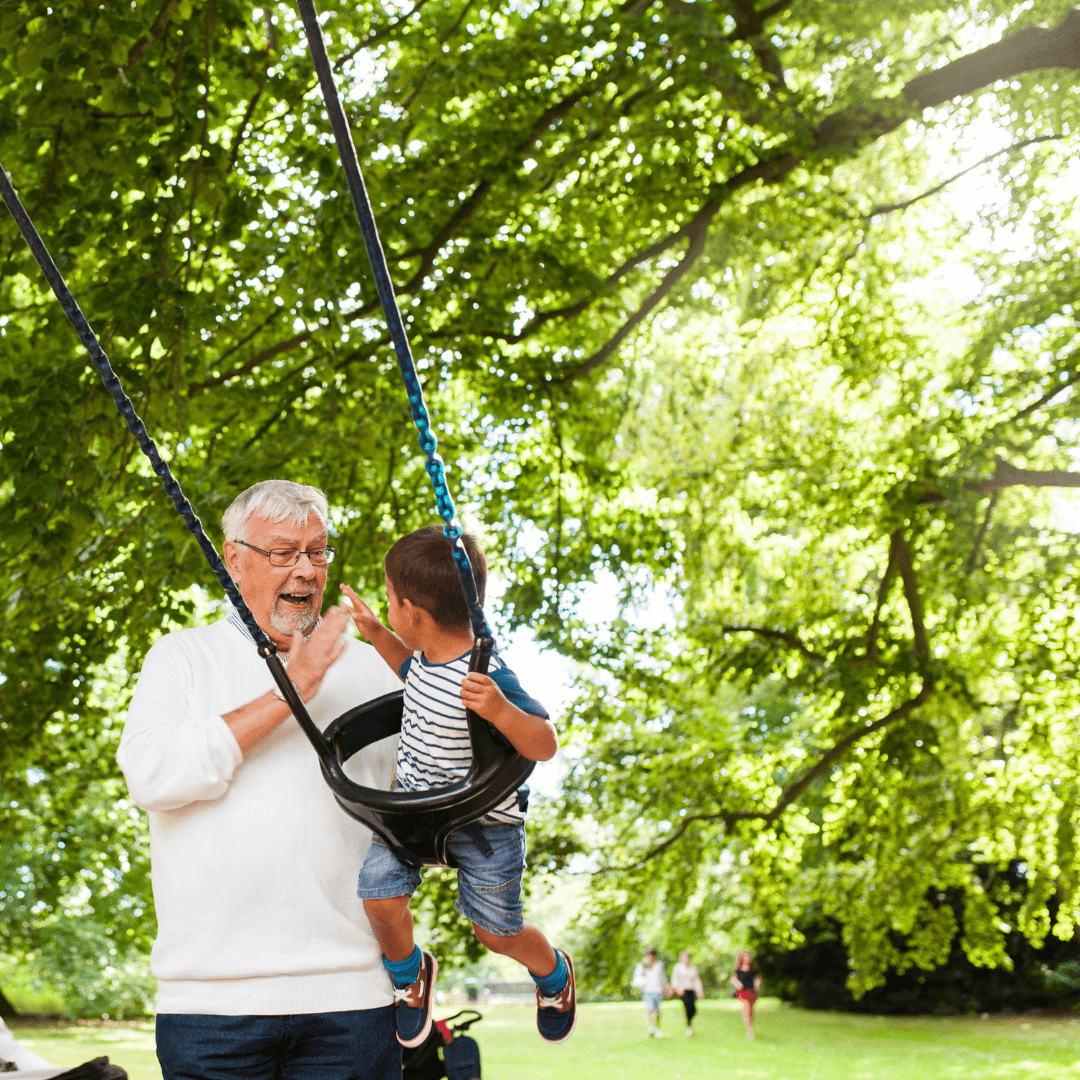 An elderly man playfully pushes a child on a swing in a sunny, green park while others walk in the background. - Home Instead